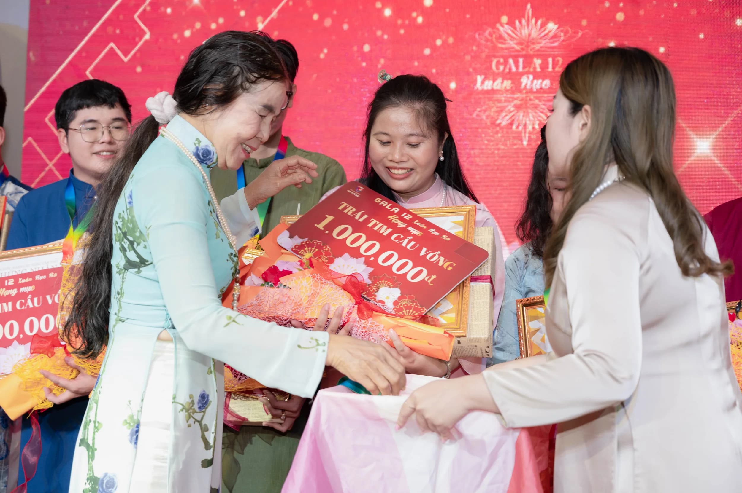 Nhiều người đang trao quà và bảng kỷ niệm trên sân khấu có phông nền đỏ chữ GALA 12 Xuân Hạo.